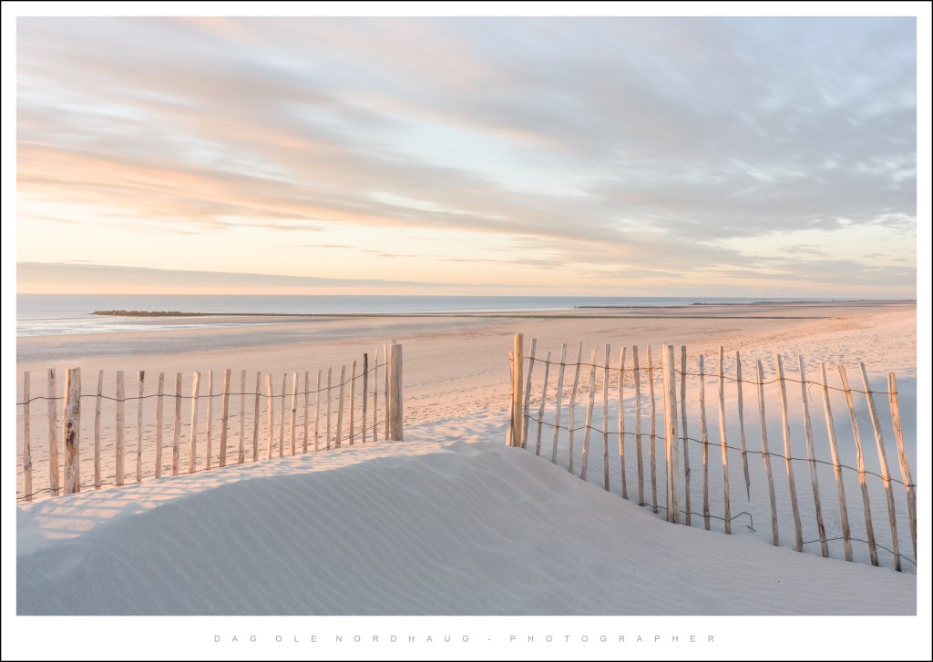 A tranquil beach scene at sunrise, featuring a wooden fence along the sandy shore and a calm ocean in the background, with soft pastel clouds in the sky.