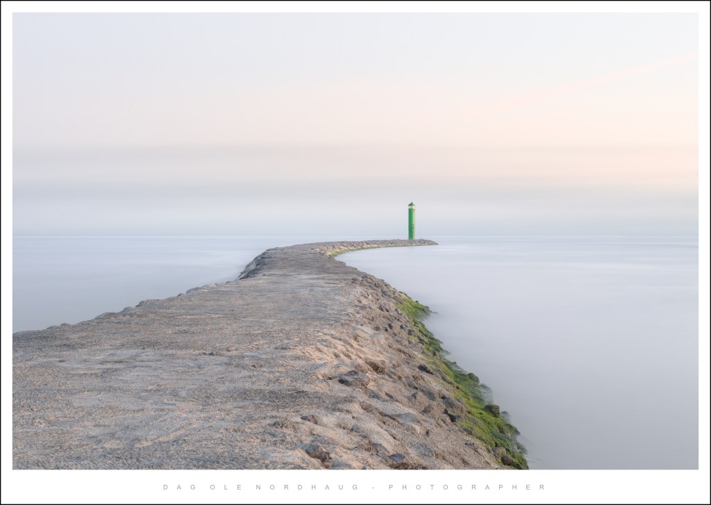 A rocky jetty extending into a calm sea, with a green lighthouse standing at the end under a soft pastel sky.