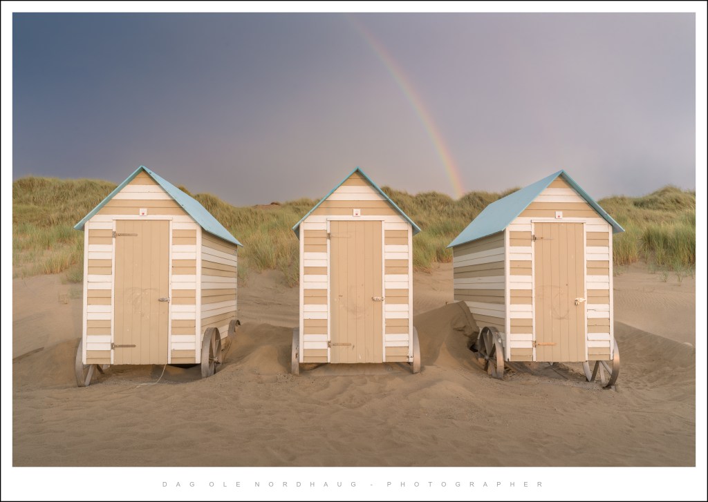 Three colorful beach huts on wheels situated on sandy terrain, with a rainbow visible in the background.