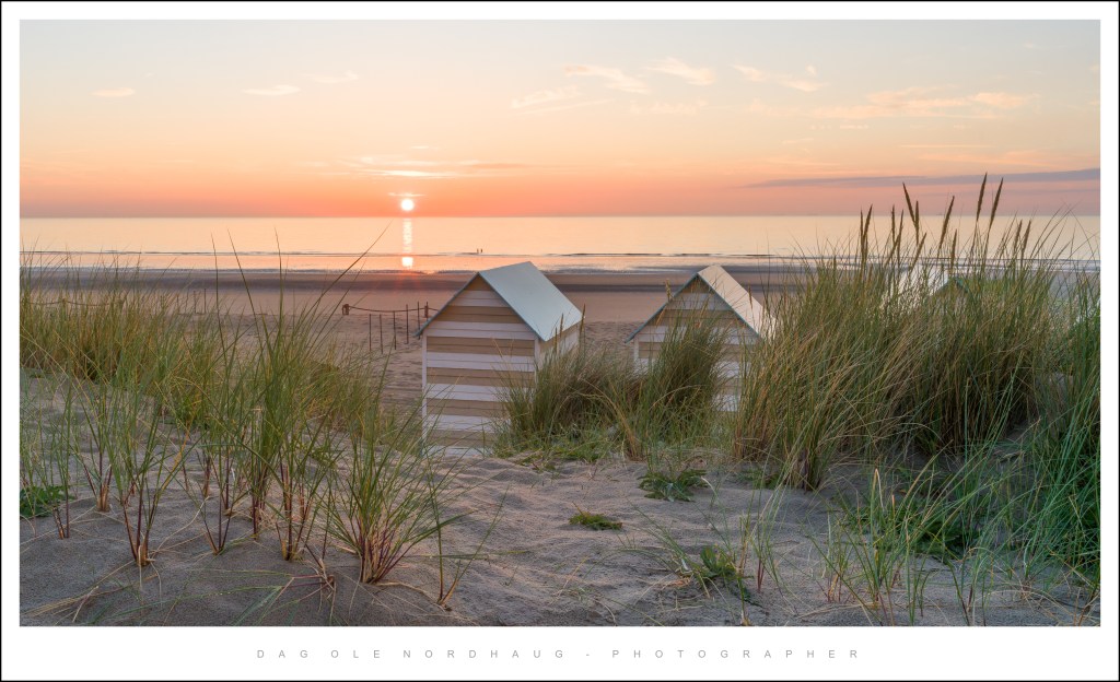 A serene beach scene at sunset featuring small beach huts surrounded by tall grass and soft sand, with calm waters and a colorful sky.