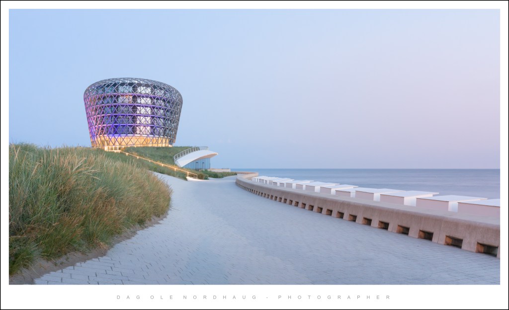 Modern architectural building with a uniquely shaped glass exterior, situated by the seaside along a paved walkway and grassy area.
