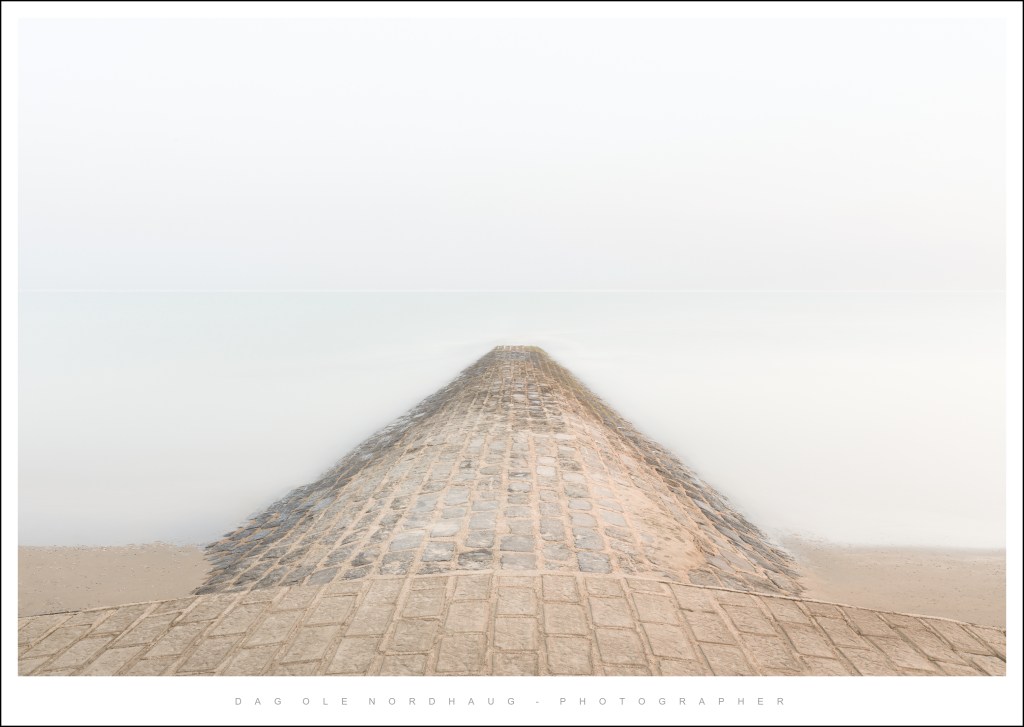 A long, stone jetty extending into a calm body of water, surrounded by foggy weather.