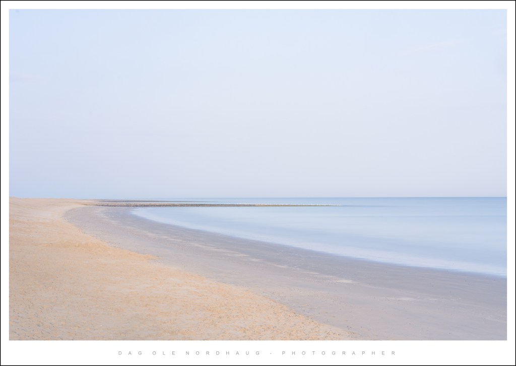 A serene beach scene with soft sand curving along the shore, meeting calm, reflective waters under a pale sky.