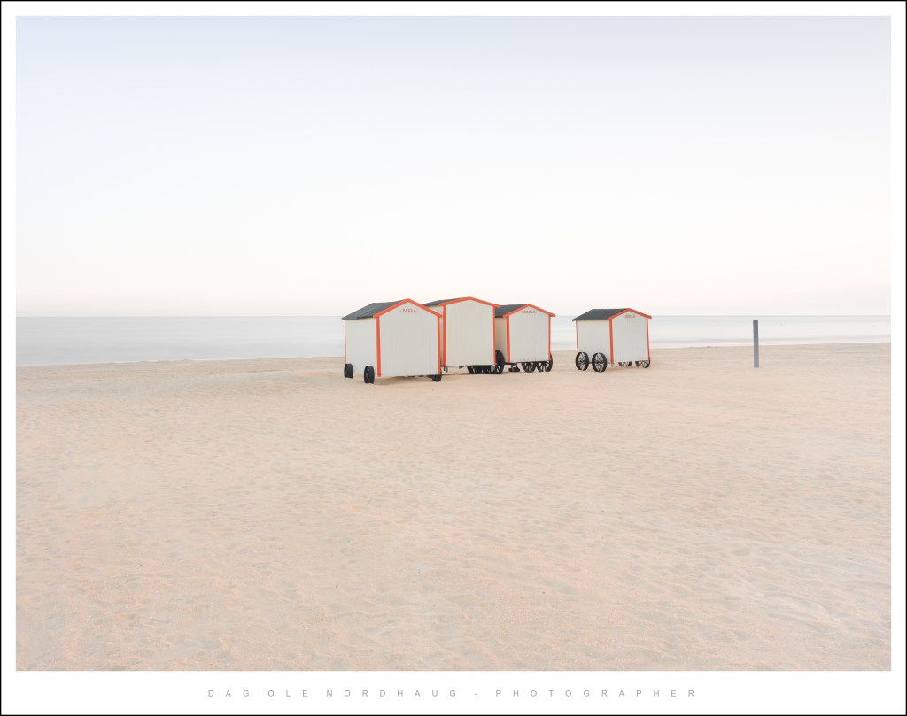 Beach scene featuring four colorful beach huts on a sandy shore with a calm ocean in the background.