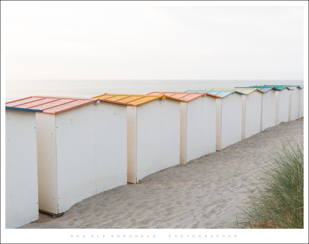 A row of colorful beach huts with slanted roofs, located on a sandy beach near the ocean, under a light overcast sky.