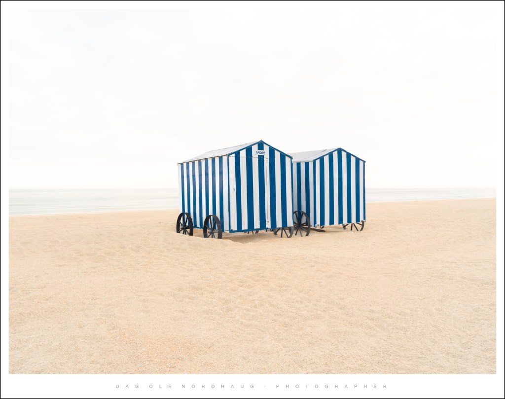 Two blue and white striped beach huts on a sandy beach with calm waters in the background.