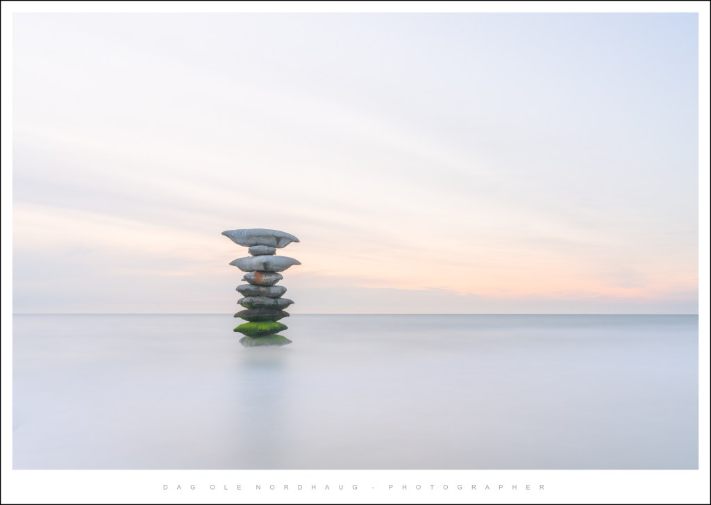 A stack of smooth stones balanced on top of each other, set against a calm ocean and soft pastel sky during sunset.
