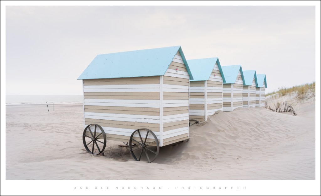 A row of beach huts with light blue roofs and white wooden walls, located on a sandy beach with the ocean in the background.