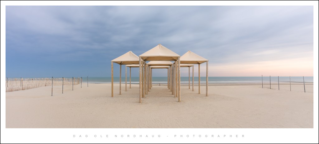 Beach scene featuring several wooden structures with fabric roofs against a cloudy sky and ocean in the background.